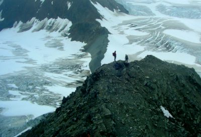 Two hikers stand on a tall moraine overlooking two glaciers in the Alaskan backcountry.
