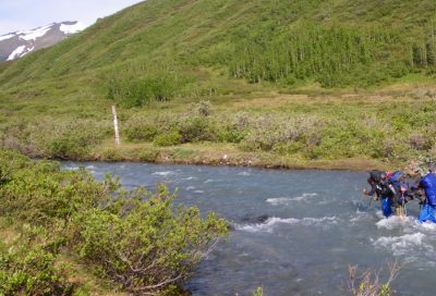 Two hikers wade through a cold river during a backpacking trip in Alaska.