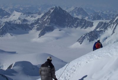 Two mountaineers ascending Mt. Fairweather with sweeping glacier valleys and rugged peaks below.