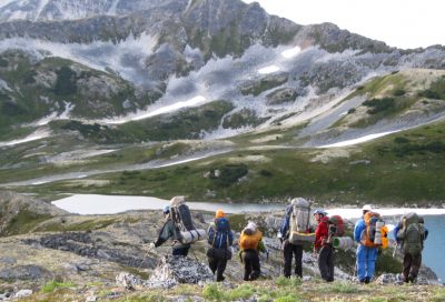 Hikers making their way up a high mountain pass on Mt. Ripinsky, overlooking dramatic alpine peaks and a turquoise glacial lake.