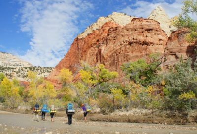 Hikers crossing a shallow stream in a scenic Utah red rock canyon with fall colors.