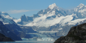 Alaska glacier flowing between rugged, snow-covered peaks on a clear day.